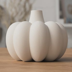 May include: A cream-colored pumpkin-shaped vase with a textured surface. The vase has a short, cylindrical neck and a rounded body with vertical ridges. It sits on a wooden surface.