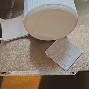May include: A white cylindrical object and a small square object with a textured surface sit on a silver-colored surface. A piece of white paper is also present. The objects appear to be related to 3D printing.