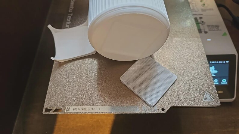 May include: A white cylindrical object and a small square object with a textured surface sit on a silver-colored surface. A piece of white paper is also present. The objects appear to be related to 3D printing.