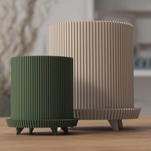 May include: Two cylindrical planters with vertical grooves. One is dark green, the other is beige. Both have a matching base with three small legs. The planters are made of wood and are on a wooden surface.