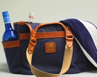 Puede incluir: Bolso tote azul marino con detalles en cuero marrón, que contiene una botella azul y una botella transparente. Una toalla a rayas azules y blancas descansa sobre el bolso. El bolso tiene una correa beige y un pequeño logotipo.