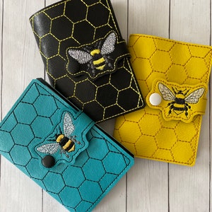 May include: Three small, colorful wallets with bee embroidery. The wallets are black, teal, and yellow, each with a honeycomb pattern. The bee design is detailed with yellow and black stripes and silver wings. Each wallet has a snap closure.
