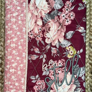 May include: A burgundy quilted fabric book cover with a floral pattern. The cover has a pink and white floral fabric border. The text "be still and know" is embroidered in gray on the cover.
