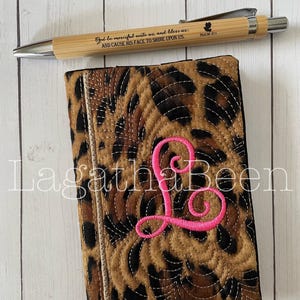 May include: A leopard print journal cover with a pink embroidered letter "L" and a matching pen. The pen is made of bamboo and has the text "God be merciful unto us, and bless us; AND CAUSE HIS FACE TO SHINE UPON US."