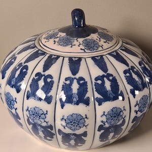 May include: A white and blue ceramic lidded jar with a rounded shape. The jar is decorated with blue floral and leaf patterns. The lid has a small, dark blue knob. The overall design is symmetrical and detailed, suggesting a decorative or storage item.