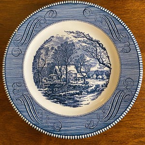 May include: A blue and white decorative plate featuring a winter landscape scene. The plate has a blue border with a textured design and decorative scrollwork. The rim is scalloped.