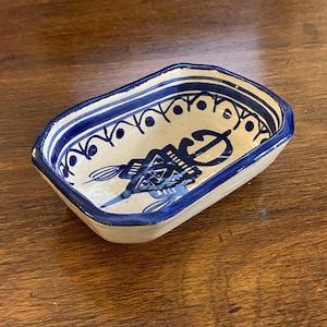 May include: A rectangular ceramic dish with a blue and white design. The rim is painted blue with small dots and curved lines. The interior features a central blue design with lines and shapes. A decorative piece.