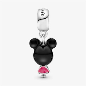 May include: A silver charm with a black Mickey Mouse head and a pink dress. The charm has the word "Minnie" engraved on the top. The charm is designed to be a pendant or a bead for a bracelet.