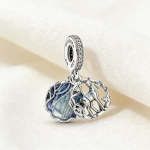 Puede incluir: Un charm de plata con un ciervo dentro de una corona y un diseño de árbol azul y plateado. El charm tiene un pequeño lazo con la inscripción "PANDORA" y "ALE S925". El charm está sobre un fondo blanco.