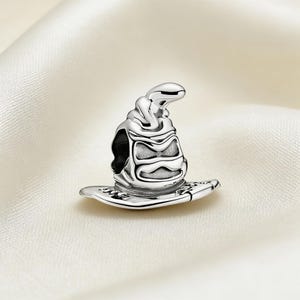 May include: A silver charm in the shape of a wizard's sorting hat. The hat has a detailed design with a wide brim and a textured surface. The charm is likely made of sterling silver or a similar metal.
