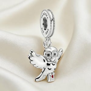 Puede incluir: Un charm de plata con un b&uacute;ho con las alas extendidas, sosteniendo un peque&ntilde;o paquete con un coraz&oacute;n rojo. El charm tiene una parte superior circular con la palabra "PANDORA" y "ALE S925" grabadas. El charm de b&uacute;ho est&aacute; suspendido de la parte superior circular.