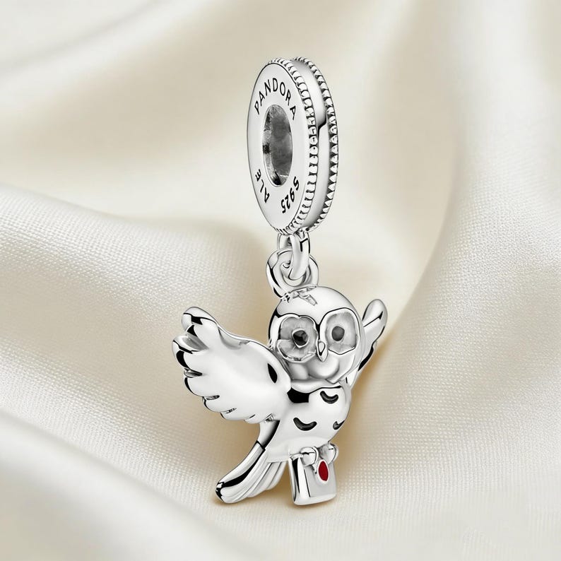 Puede incluir: Un charm de plata con un b&uacute;ho con las alas extendidas, sosteniendo un peque&ntilde;o paquete con un coraz&oacute;n rojo. El charm tiene una parte superior circular con la palabra "PANDORA" y "ALE S925" grabadas. El charm de b&uacute;ho est&aacute; suspendido de la parte superior circular.