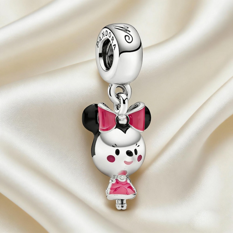 May include: A silver charm featuring a cartoon character with black ears, a pink bow, and a pink dress. The charm has a round top with the word "Pandora" and a small loop for attaching to a bracelet or necklace.