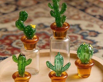 May include: A collection of five glass cactus figurines in amber-colored pots. Two cacti are attached to clear glass bottles, while three are standalone. Each green cactus features small yellow or white details, arranged on a round wooden tray.