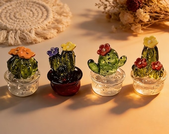 May include: Four glass cactus figurines with colorful flower accents. Each cactus is a different shape and color, with orange, purple, red, and yellow flowers. The cacti sit in clear glass pots, creating a decorative display.