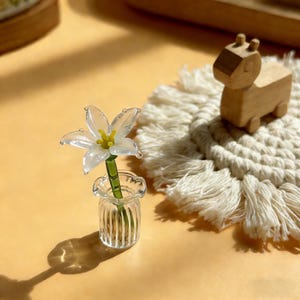 May include: A clear glass flower in a small vase with a green stem and yellow center. A wooden animal figurine sits on a white macrame coaster. The objects are on a yellow surface.