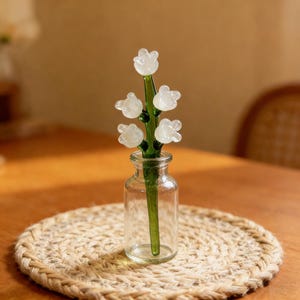 May include: A clear glass vase holds a delicate glass flower arrangement. The arrangement features a green stem with six white, translucent flower blooms. The vase sits on a round, woven placemat, creating a simple, elegant display.