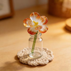 May include: A miniature glass flower in a small clear glass bottle, resting on a beige crocheted coaster. The flower has orange and white petals with a yellow center. The stem is green.