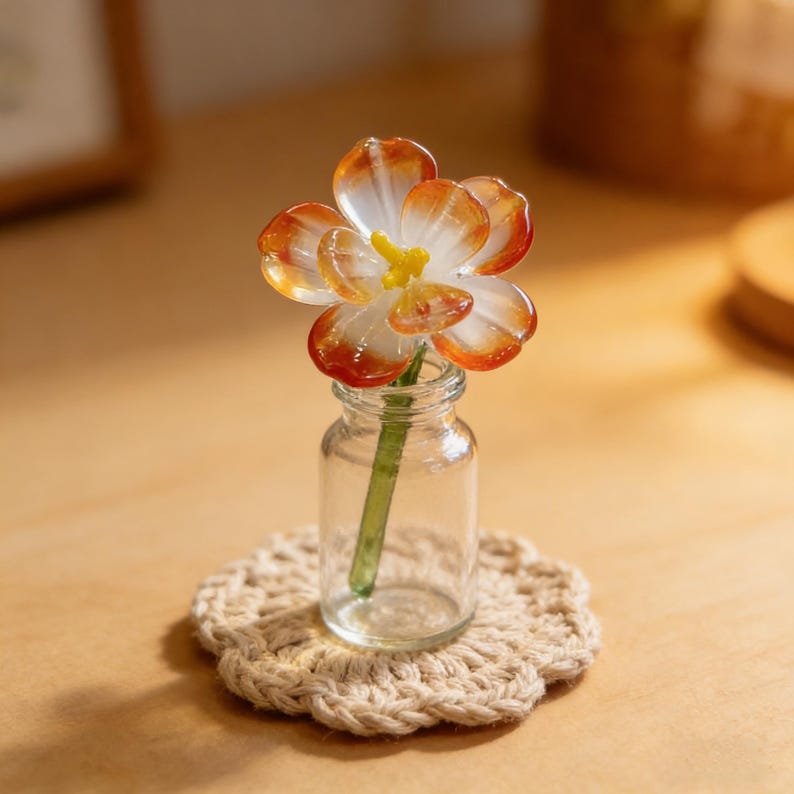 May include: A miniature glass flower in a small clear glass bottle, resting on a beige crocheted coaster. The flower has orange and white petals with a yellow center. The stem is green.