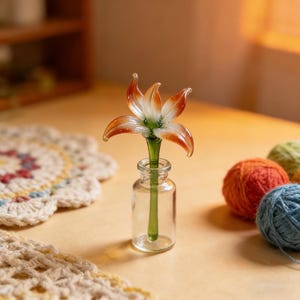 May include: A clear glass vase holds a delicate, handcrafted glass flower. The flower has white petals with orange tips and a green stem. The vase sits on a light brown surface, with colorful yarn balls in the background.