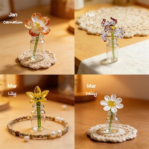 May include: Four glass flower sculptures in small glass bottles. The flowers are a carnation, iris, lily, and daisy. Each bottle sits on a decorative coaster or is surrounded by a bracelet. The text "Jan carnation", "Feb iris", "Mar lily", and "Mar daisy" is visible.