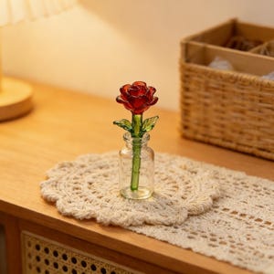 May include: A miniature glass rose in a small clear glass vase. The rose is a deep red color with green leaves and stem. The vase sits on a cream-colored, crocheted doily, with a wooden cabinet and basket in the background.