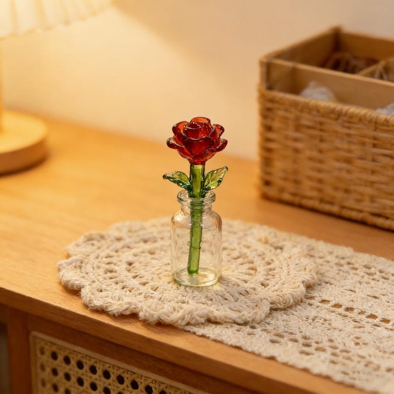 May include: A miniature glass rose in a small clear glass vase. The rose is a deep red color with green leaves and stem. The vase sits on a cream-colored, crocheted doily, with a wooden cabinet and basket in the background.