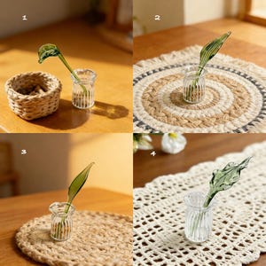 May include: A clear glass bud vase with a green glass leaf stem. The vase is small and ribbed, and the leaf has detailed veining. The vase is displayed on a wooden table with a woven basket and a woven placemat.