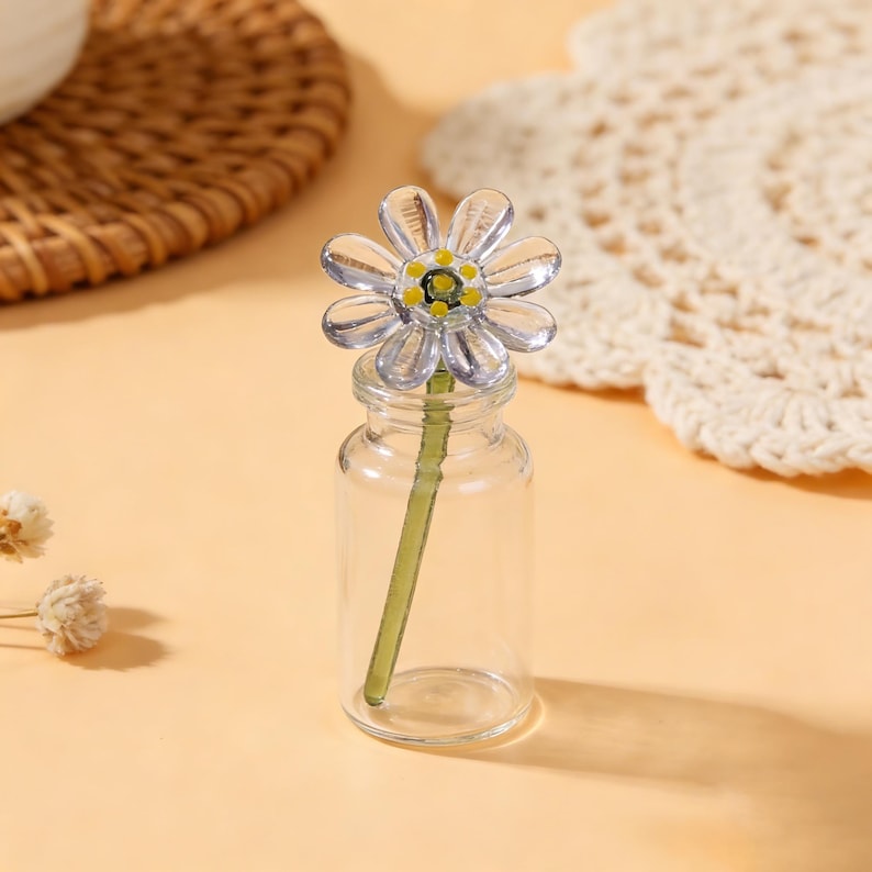 May include: A clear glass bud vase with a daisy-shaped flower. The flower has clear petals with yellow and green accents. The vase is cylindrical and sits on a light orange surface. A woven basket and doily are in the background.