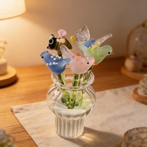 May include: A clear glass vase holds a collection of colorful glass figurines. The figurines include birds, a bee, and a butterfly, each with intricate details and vibrant colors. The vase sits on a white cloth.