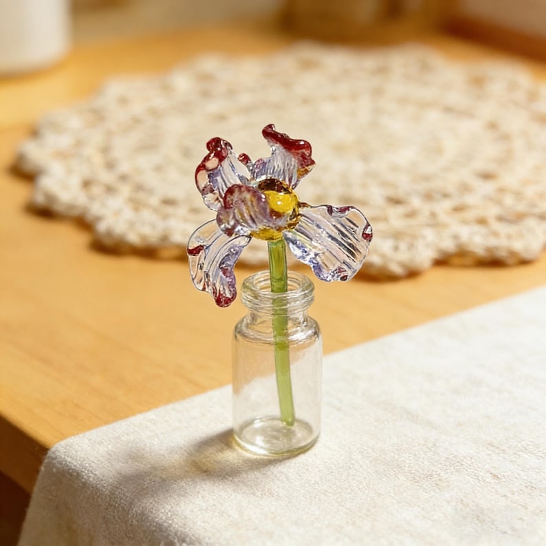May include: A clear glass iris flower with red and yellow accents in a small clear glass bottle. The flower has delicate petals and a green stem. The bottle sits on a white cloth.