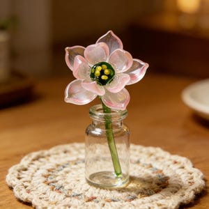 May include: A delicate glass flower with pink petals and a green and yellow center is displayed in a small, clear glass bottle. The flower sits on a beige, crocheted coaster, creating a charming, decorative arrangement.