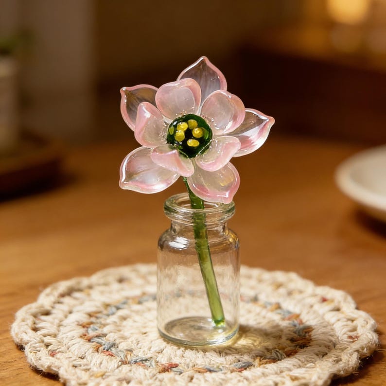 May include: A delicate glass flower with pink petals and a green and yellow center is displayed in a small, clear glass bottle. The flower sits on a beige, crocheted coaster, creating a charming, decorative arrangement.