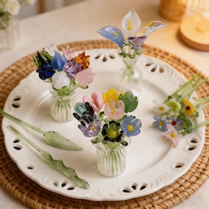 May include: Several colourful glass flower bouquets in small, clear vases, displayed on a white plate with a scalloped edge. The flowers feature blue, pink, yellow, and green petals. Two green glass stems are also visible.