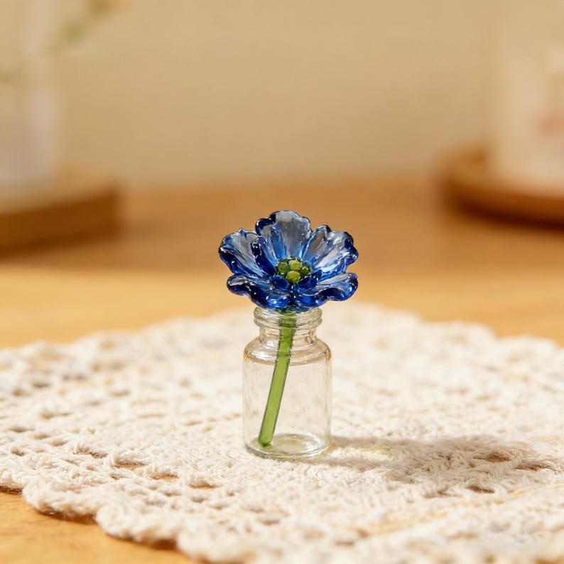 May include: A small, clear glass bottle holds a delicate blue glass flower with a green stem. The flower sits on a cream-colored, crocheted doily. The background is a soft, blurred neutral tone.