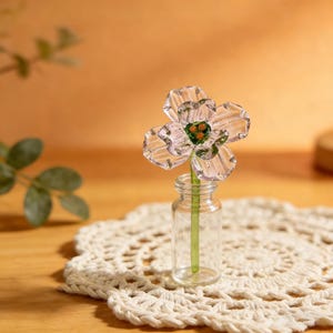 May include: A clear glass flower with a green stem is displayed in a small glass bottle. The flower has five petals with a green and orange center. The bottle sits on a white, crocheted doily. The background is a warm, neutral tone.