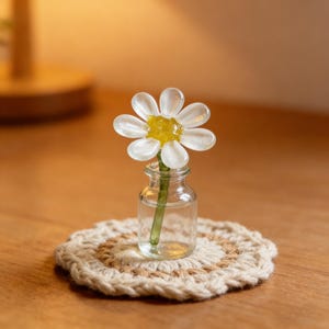 May include: A glass daisy flower in a small clear glass bottle, with a green stem, resting on a beige and brown crocheted coaster. The daisy has white petals and a yellow center. The items are on a wooden surface.