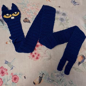 Puede incluir: Una bufanda azul de ganchillo hecha a mano con un diseño de cara de gato. La cara del gato tiene ojos amarillos y detalles blancos. La bufanda está hecha de un hilo suave y texturizado y se muestra sobre una tela con estampado floral.