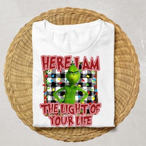 May include: White t-shirt featuring a green cartoon character with the text "HERE I AM THE LIGHT OF YOUR LIFE" in red. The character is set against a black and white plaid background with colorful string lights.