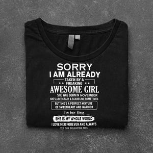 Pode incluir: Camiseta preta com texto branco. O texto diz "SORRY I AM ALREADY TAKEN BY A FREAKING AWESOME GIRL" e continua com texto adicional sobre a garota, incluindo "I'm her king" e "SHE IS MY WHOLE WORLD."