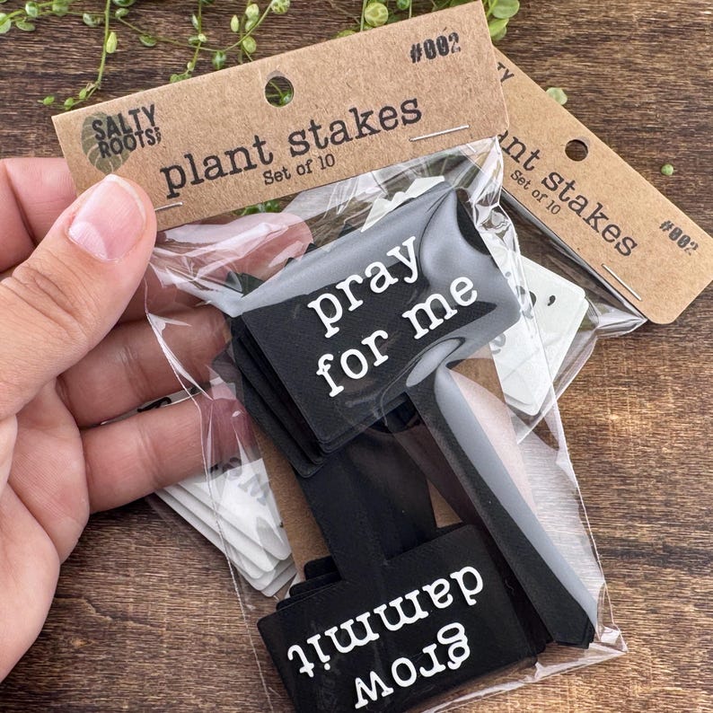 May include: A set of ten black plant stakes in a clear plastic bag with a brown cardboard header labeled Salty Roots plant stakes set of 10 #002. The stakes feature white text including the phrases pray for me and grow dammit.