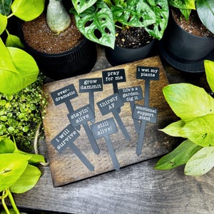 May include: A set of nine black plant markers with white text featuring phrases including "overdramatic," "grow dammit," "pray for me," "I wet my plants," "stayin' alive," "thirsty AF," "life's a garden. dig it," "emotional support plant," and "i will survive."
