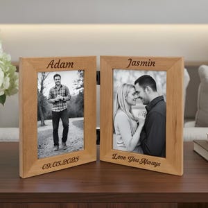 Op de afbeelding: Een houten dubbele fotolijst met twee zwart-witfoto's. Het linkerframe heeft de naam "Adam" en de datum "09.05.2025" gegraveerd. Het rechterframe heeft de naam "Jasmin" en de tekst "Love You Always" gegraveerd.