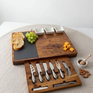 Op de afbeelding: Een houten kaasplankenset met een leistenen inzet, kleine witte kommen en kaasmesjes. Inclusief druiven, brood, kaasblokjes, honing en amandelen. De plank is gegraveerd met "Rachel & Brad".