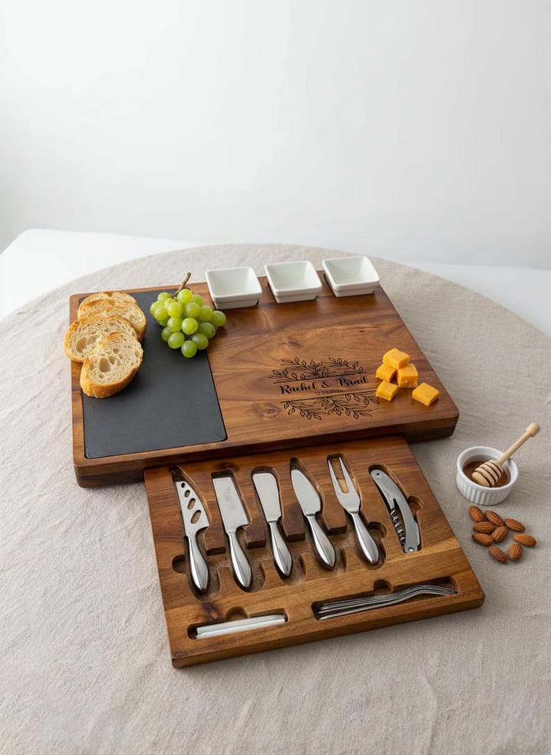 Op de afbeelding: Een houten kaasplankenset met een leistenen inzet, kleine witte kommen en kaasmesjes. Inclusief druiven, brood, kaasblokjes, honing en amandelen. De plank is gegraveerd met "Rachel & Brad".