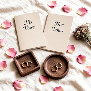 Two vow books labeled His Vows and Her Vows lie above a square and a round engraved wooden ring dish holding gold rings, scattered among rose petals and small purple flowers.