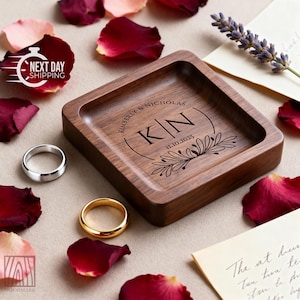 A square wooden ring dish is engraved with the initials K N and wedding details, surrounded by red rose petals, a lavender sprig, and two wedding rings.