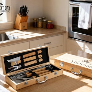 May include: A wooden barbecue tool set in a rectangular carrying case with metal latches and a handle. The lid is engraved with the name Thomas and a flame icon. Inside, stainless steel tools with wooden handles are secured by black elastic straps.
