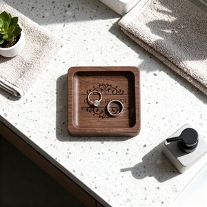 A square wooden ring dish with a custom floral engraving holds a diamond ring and a plain band, set on a speckled countertop between two folded towels and a soap dispenser.