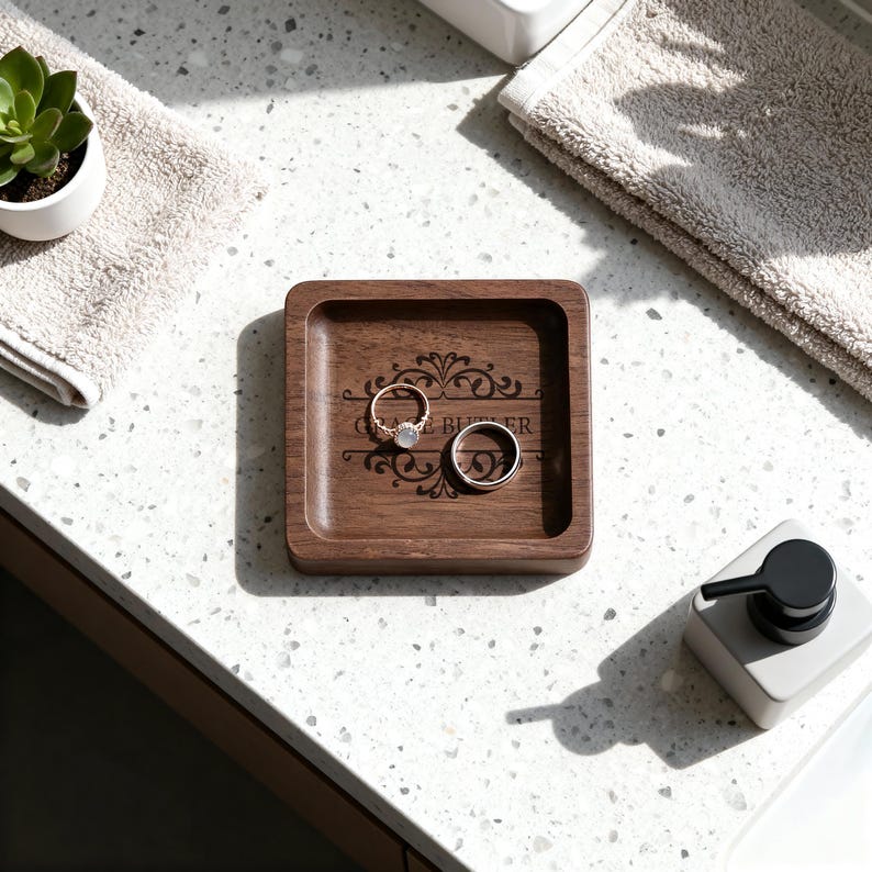 A square wooden ring dish with a custom floral engraving holds a diamond ring and a plain band, set on a speckled countertop between two folded towels and a soap dispenser.